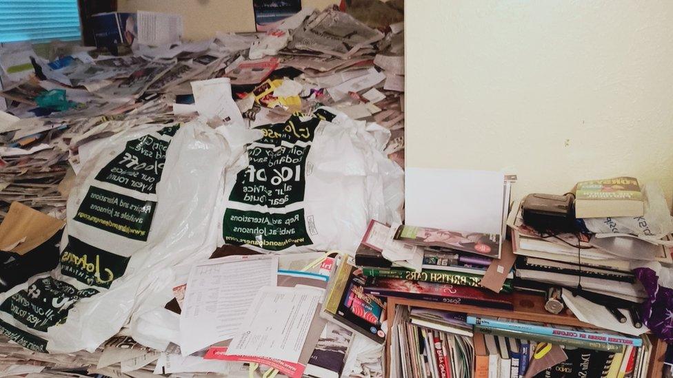 A bedroom full of hoarded items and old papers. The bed is not visible and each surface is covered with piles of items