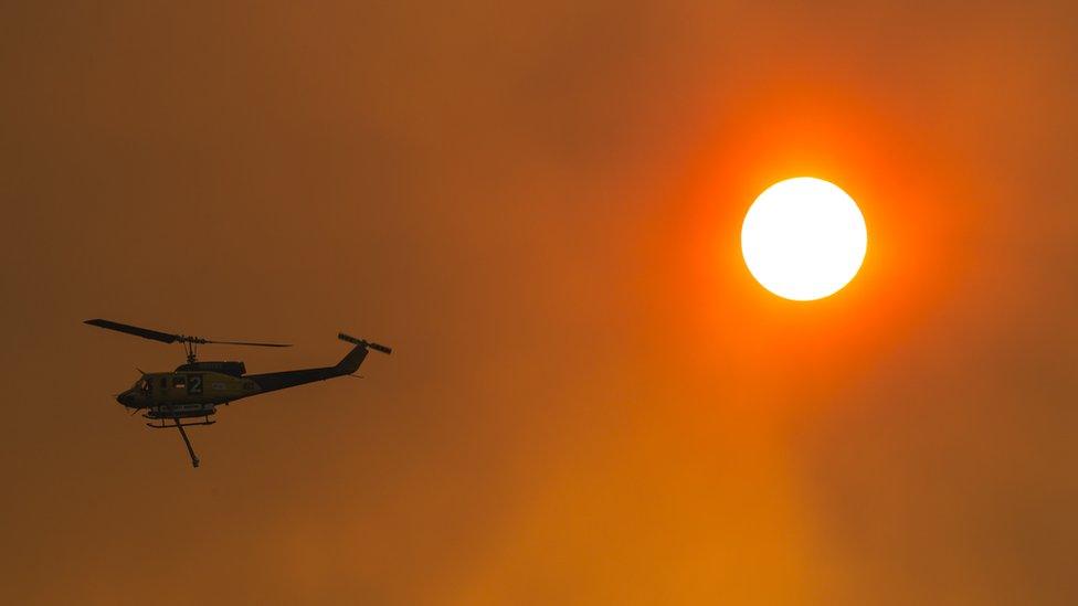 A water-bombing helicopter silhouetted on a red, smoke-filled sky next to the sun