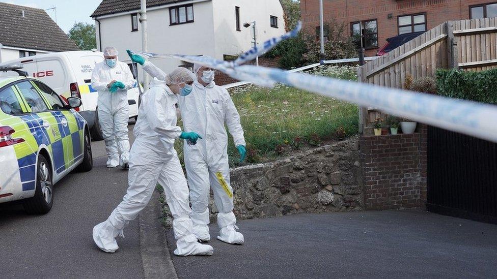 Officers in white protective clothing walk under police tape on driveway