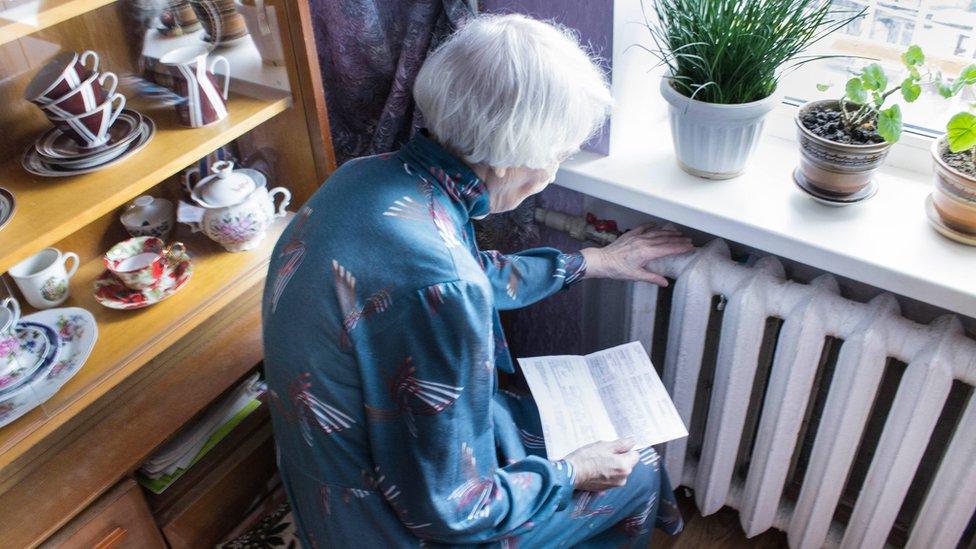 The senior woman holding gas bill in front of heating radiator.