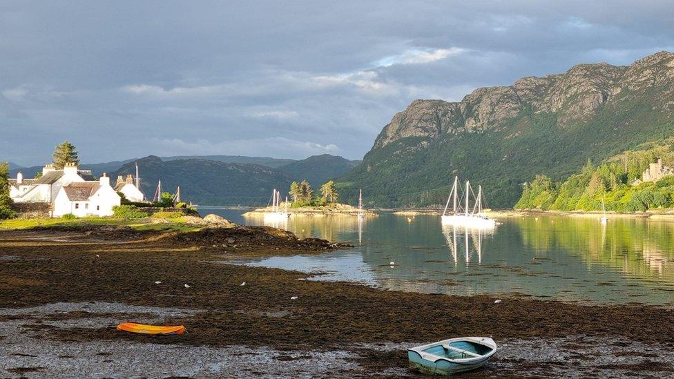 Grey rain clouds fill the sky above mountains to the right, water with boats in the centre and white houses to the right. The foreshore is muddy and wet but the sun is casting a bright glow off the houses, boats and grass