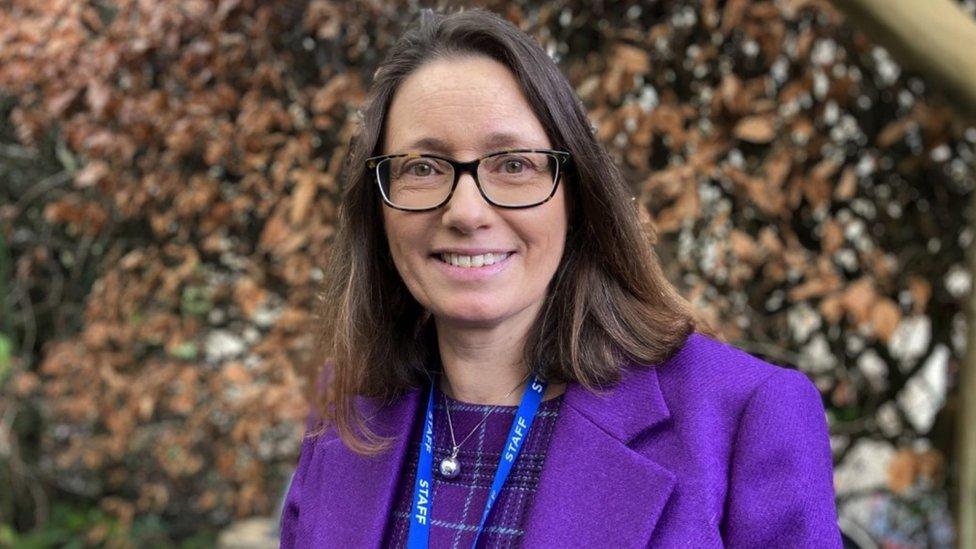 Lucy Chandler. She has dark hair and is wearing a bright purple coat. She is pictured outdoors on the school grounds, by a hedge. She is looking directly at the camera and smiling.