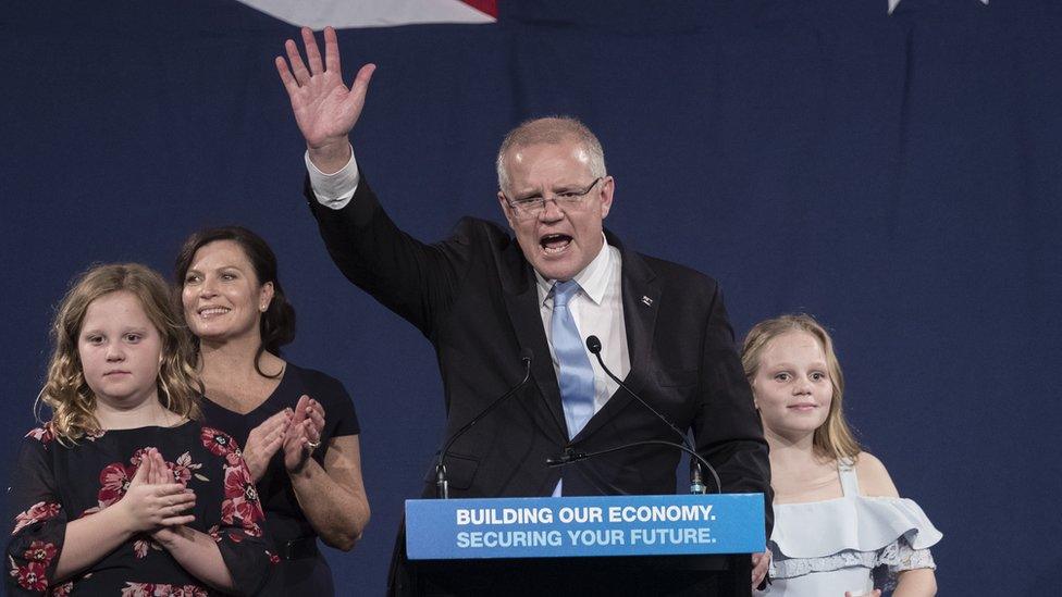 Scott Morrison waves to crowd from a podium with his wife and two daughters