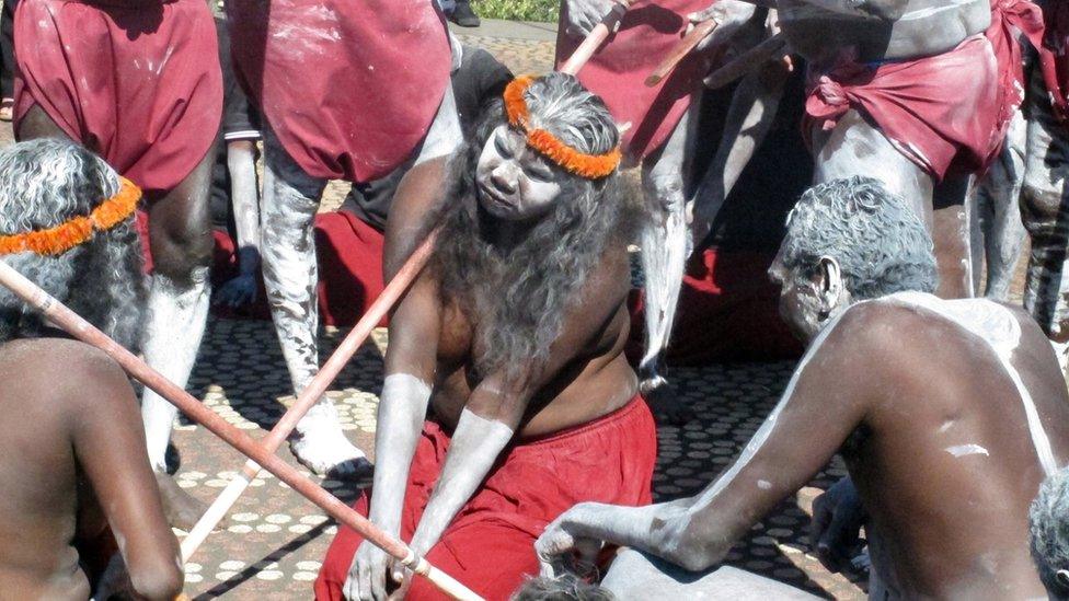 A woman kneels as Aboriginal traditional dancers carrying clap sticks and spears perform in front of Parliament House in Canberra, Australia