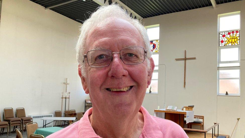 Churchwarden inside church with a cross on the wall and some stained glass in the windows