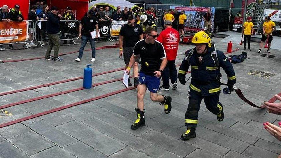 Man in firefighter uniform running whilst dragging a hose behind him, A man is running beside him.