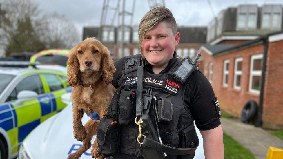 Woman in police uniform holding a small brown dog next to a police car