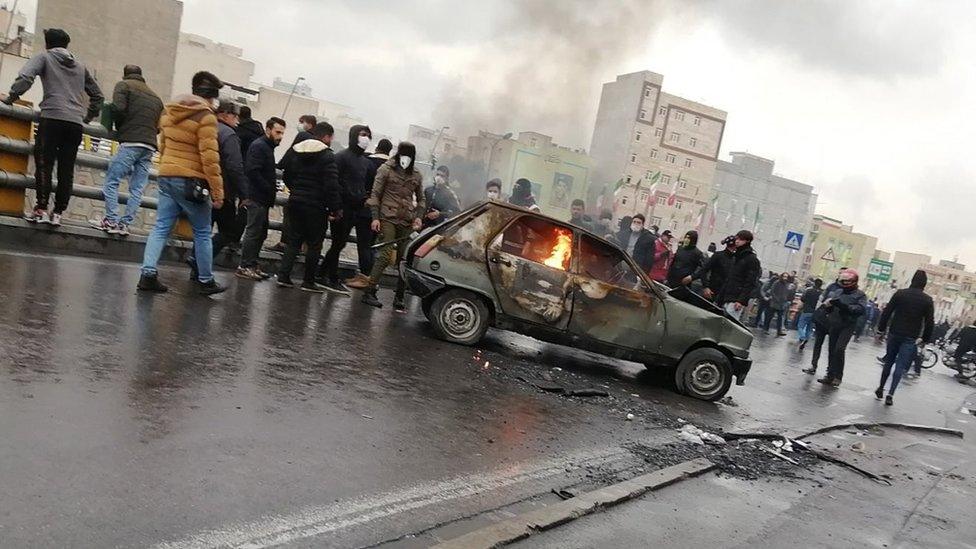 Protesters stand around a car that has been set on fire in Tehran