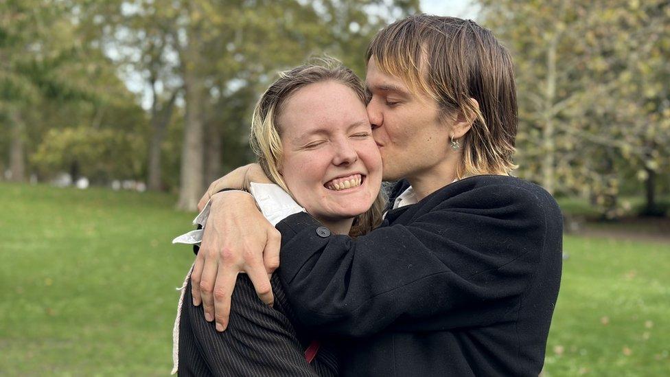 A boyfriend and girlfriend are in a park, stood on the grass, with trees behind them. The man is seen side-on. He has longish, blonde-brown hair, wears a black top and has a single hooped earring. He's embracing the woman and kissing her on the cheek. She's also got blonde-brown hair and wears a black top with white pinstripes. Her eyes are closed and she's smiling as the man squeezes her close.