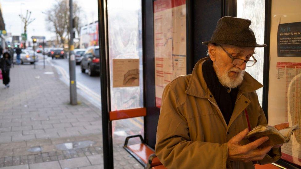 Elderly man wearing a hat and glasses reads a book at a bus stop