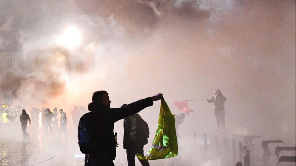 A protester holds a yellow vest a smoke billows over during a "Yellow Vest" anti-government demonstration outside the city hall of Bordeaux, south-western France, on 12 January 2019