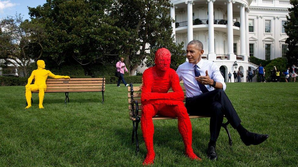 Obama sits on a bench on the White House lawn, apparently speaking to man made entirely out of red Lego bricks. The Lego man is leaning forward intently with elbows on knees.