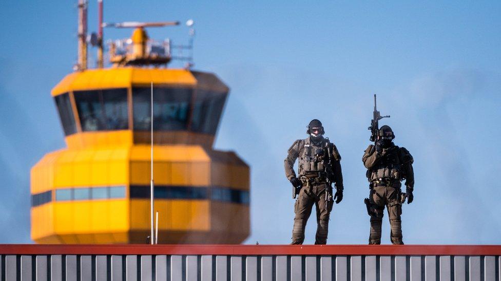 Two heavily armed and armoured officers stand on a rooftop, outlined against a clear blue sky