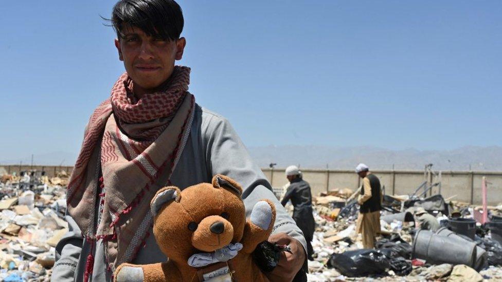 In this picture taken on June 17, 2021, a man holds a teddy bear as people look for useable items at a junkyard near the Bagram Air Base in Bagram