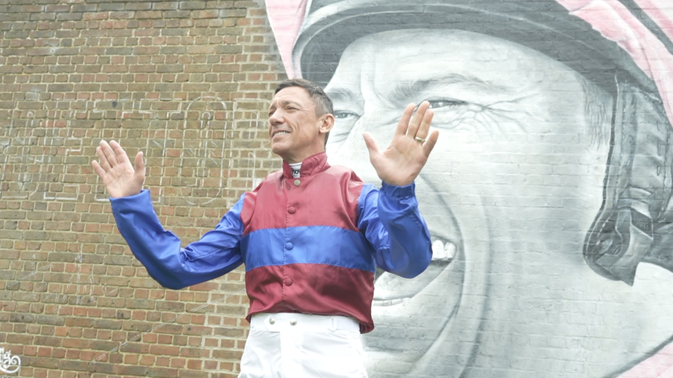 Frankie Dettori smiling in front of a monochrome mural of himself wearing a pink horseriding helmet