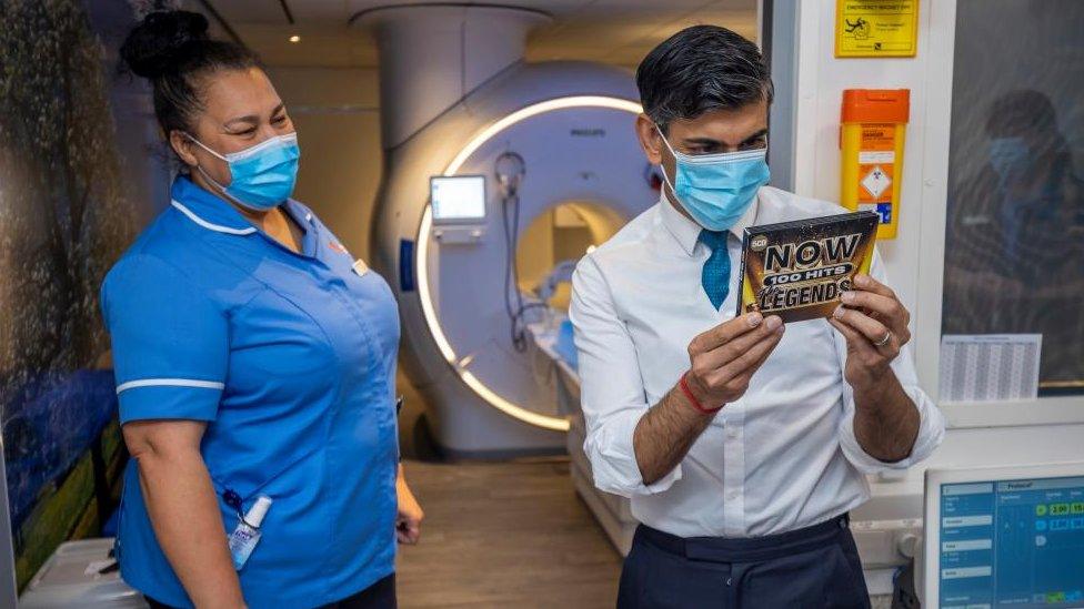 Rishi Sunak, wearing a mask, reads the back of a CD whilst a nurse looks on with an MRI scanner in the background