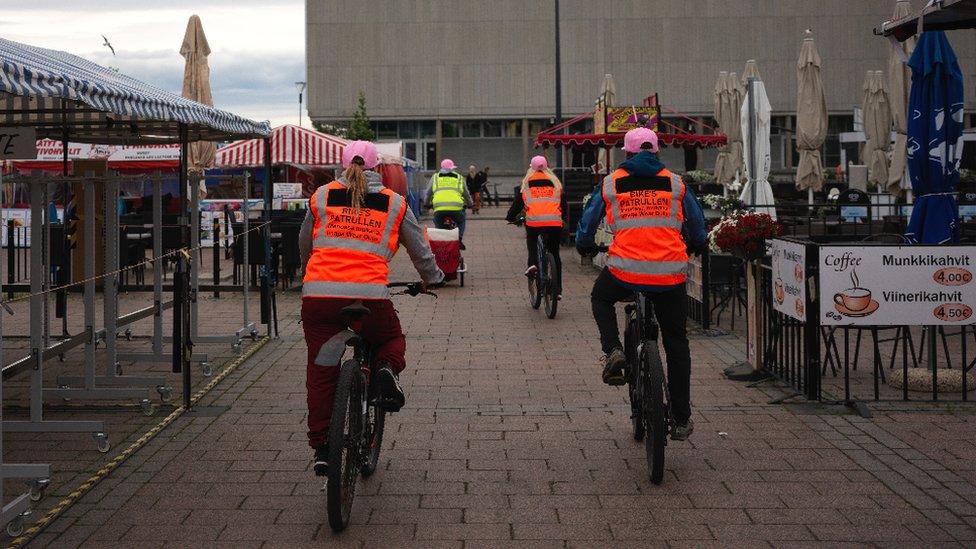 Four people in high-vis vests cycle in middle of market place and cafe area in Oulu