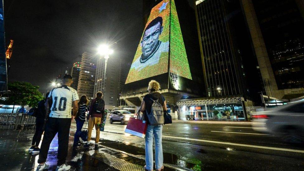 People stand on a pavement across the road from a building lit up with the face of Pelé