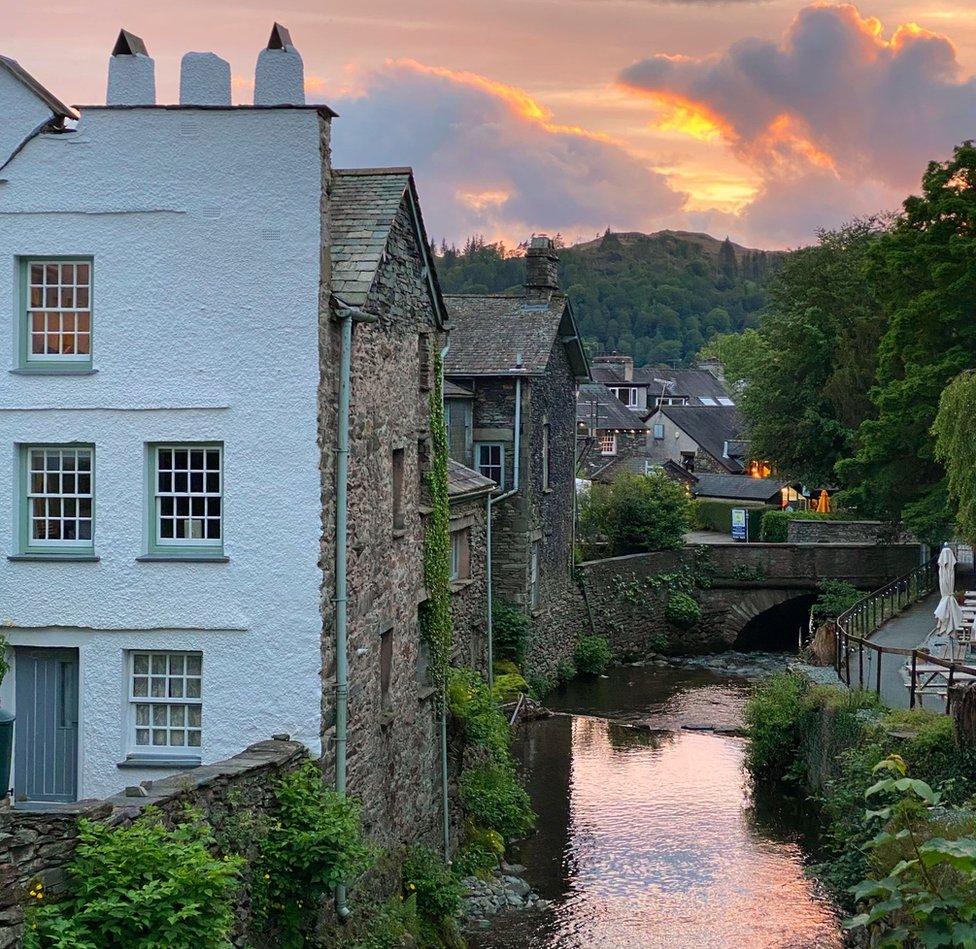 A white house made of stone stands next to a small beck reflecting a distant sunset