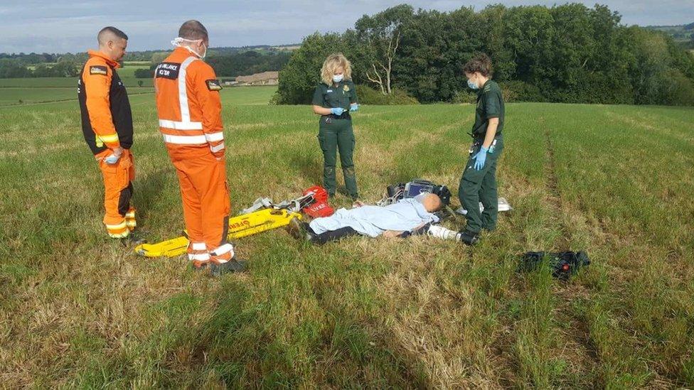 Air ambulance personnel, two in orange hi-viz and two in green paramedic clothing attend to a man lying on the floor under a blue blanket