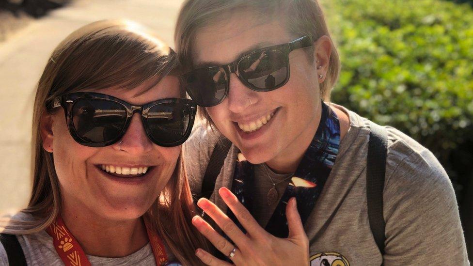 Emma (left) and Helen (right). Emma has brunette hair and is wearing black sunglasses. She is smiling at the camera. Helen has blonde hair and is standing close to Emma and is holding up her hand, showing a ring on her finger.