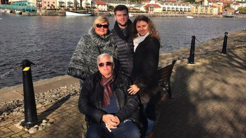 Ross Matthews pictured with his mother, father and sister. They are stood by a river with houses in the distance behind them. All four of them are smiling at the camera.