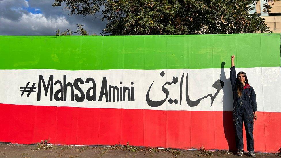 Artist Sahar Saki stands in front of her mural, with her fist closed, and raised above her head. The mural is the Iranian flag, with the words 'Mahsa Amini' written in English and Farsi