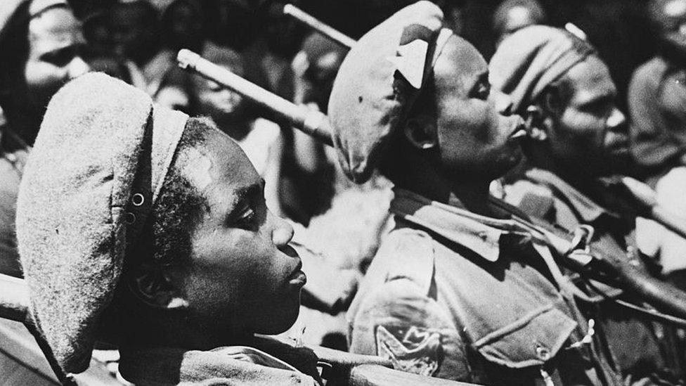 Black and white photo shows young soldiers standing at attention with guns over their shoulder