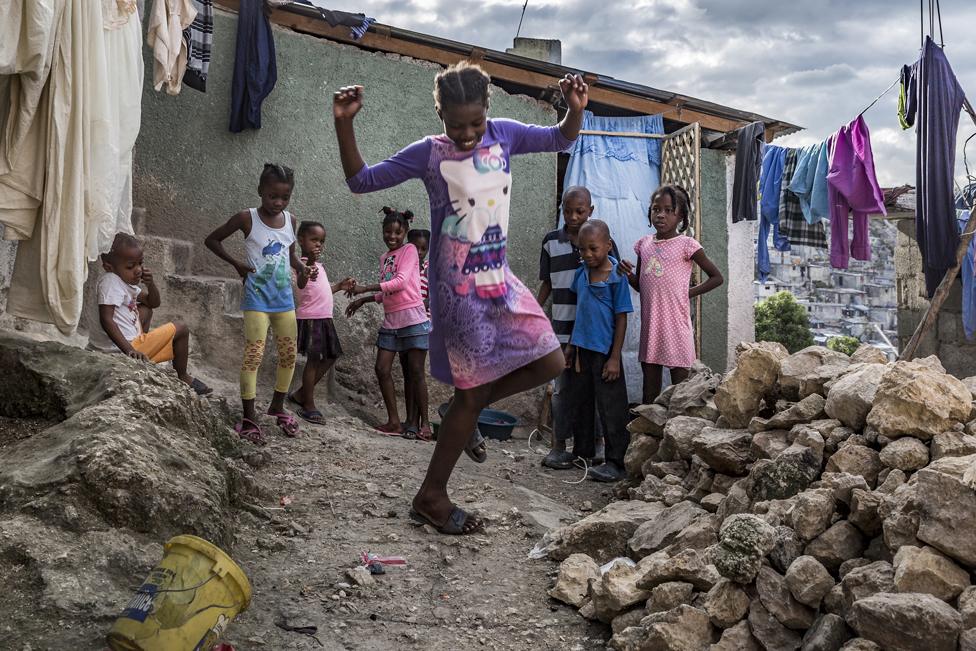 A young girl plays hopscotch in front of her house while her friends and neighbours look on