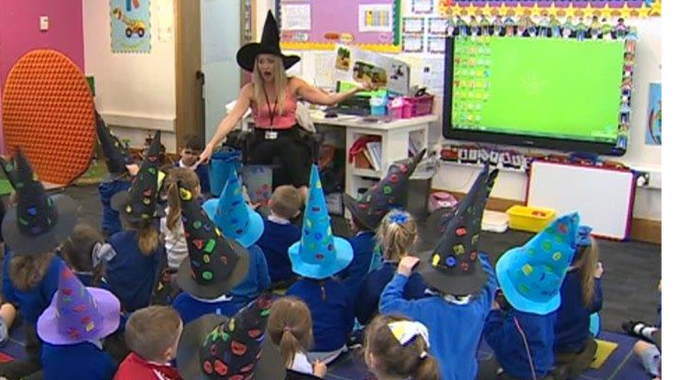 A class of children wearing witches hats listening to their teacher read a story