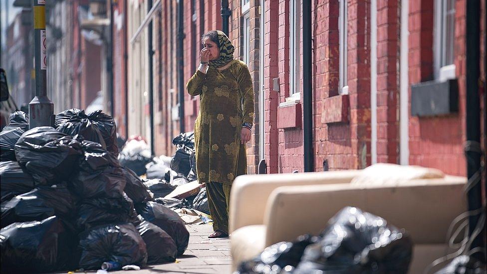 Woman in a street in Alum Rock surrounded by piles of uncollected rubbish bags