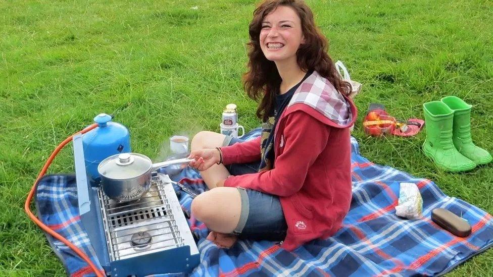 Ellie Rowe sitting on a blanket around camping gear, smiling to the camera