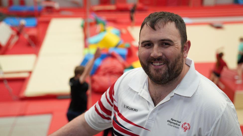 Alex Row, head coach in his coaching kit. He is standing in front of gymnastics matting and equipment.