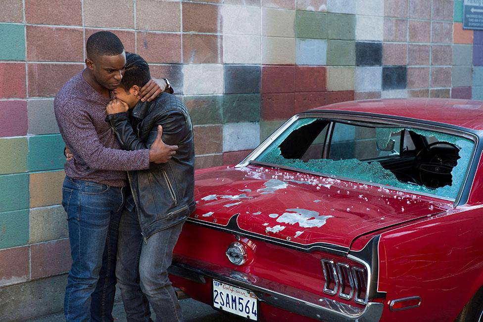 Characters Caleb and Tony hug each other in front of a smashed car