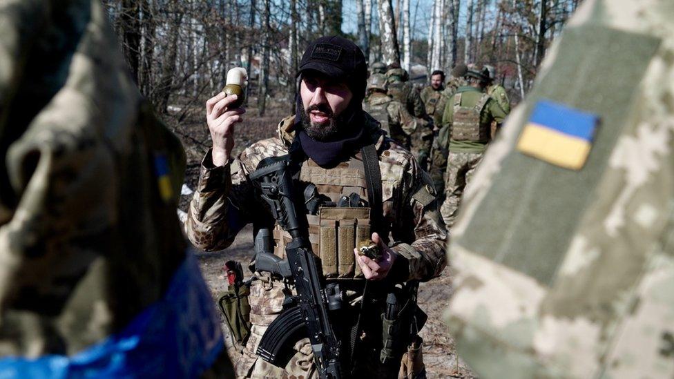 Levan holds a hand grenade up, lecturing some recruits