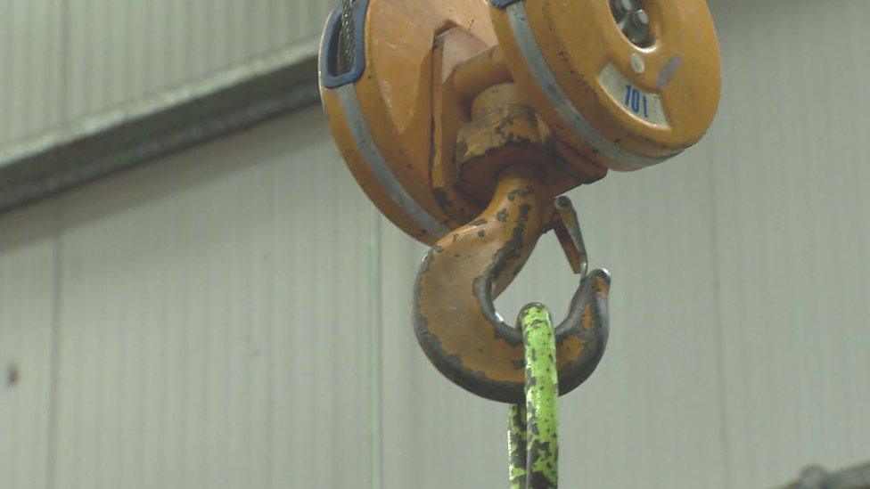 An upside down hook hanging from a piece of industrial machinery in a factory, with another piece of metal attached to it