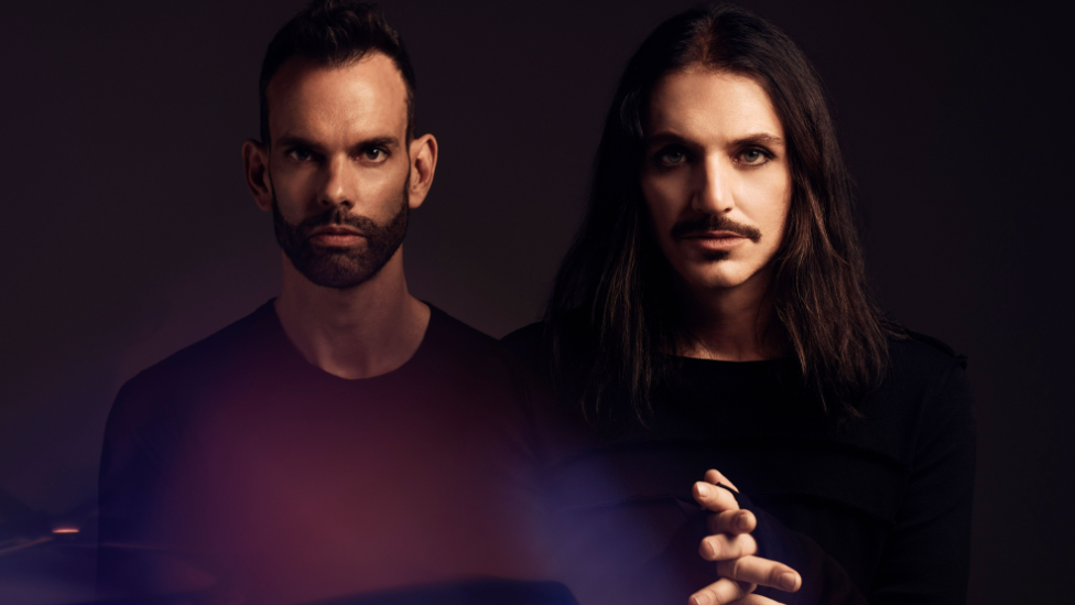 The rock band Placebo, Two men are looking at he camera, one has short dark hair and the other has long dark hair and a moustache