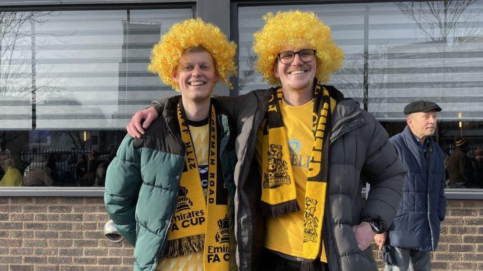 Two male Maidstone United fans wear bright yellow wigs and club kits and smile at the camera