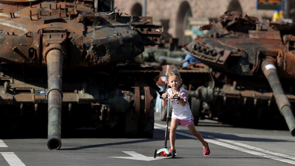 Girl on scooter in front of armoured tanks