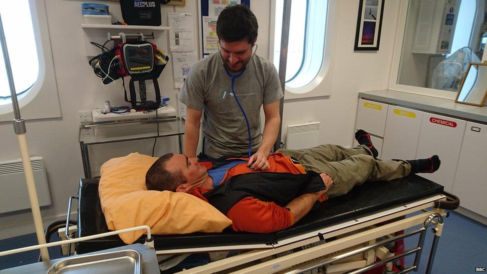 A man with a stethoscope listening to a patient's chest. The patient is laid out on a black bed. There are cupboards and medical equipment around