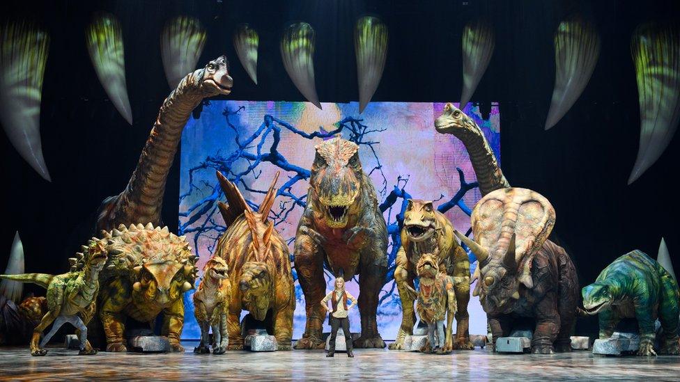 A herd of dinosaurs of different sizes on stage with Michaela Strachan stood in front of them