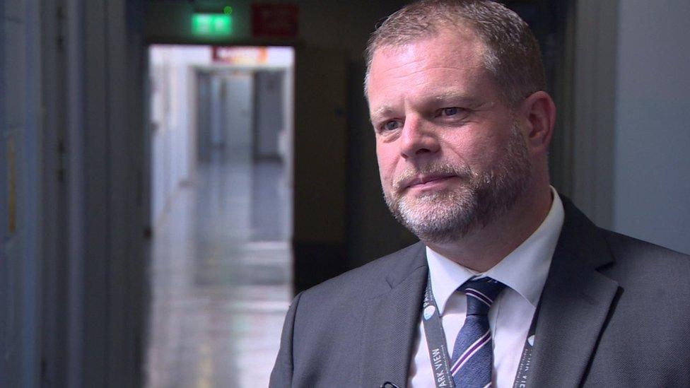 Headteacher Andy Webster wearing a grey suit and striped tie, stood in front of the corridor in the affected building