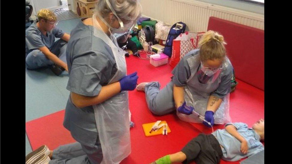 Two teachers at a special school wearing PPE masks and gowns interact with a young boy who is lying on the ground in school uniform.