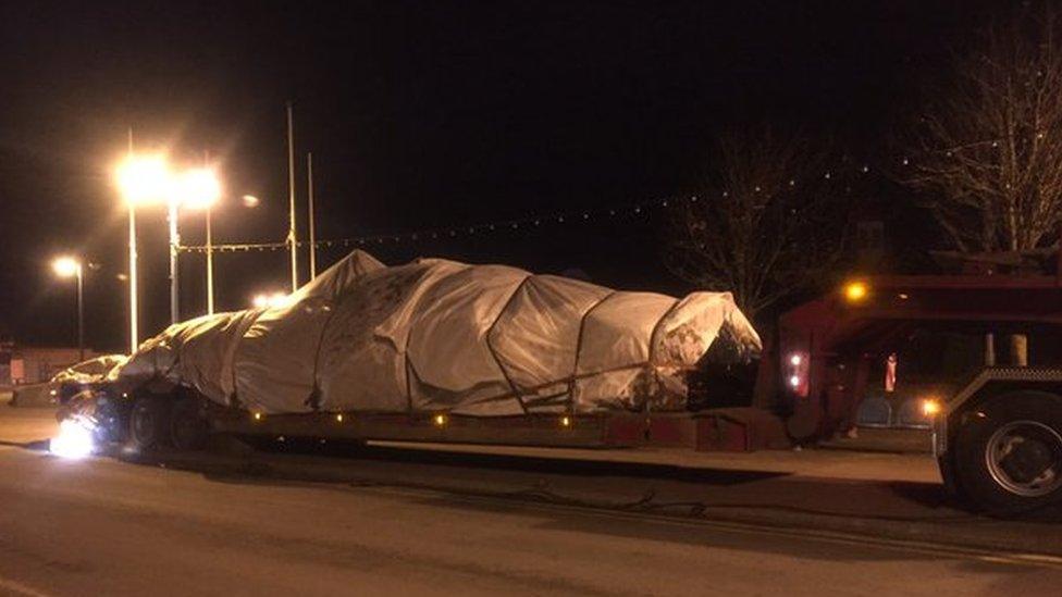 Whale wrapped up in tarpaulin loaded onto a lorry, which is travelling down a road