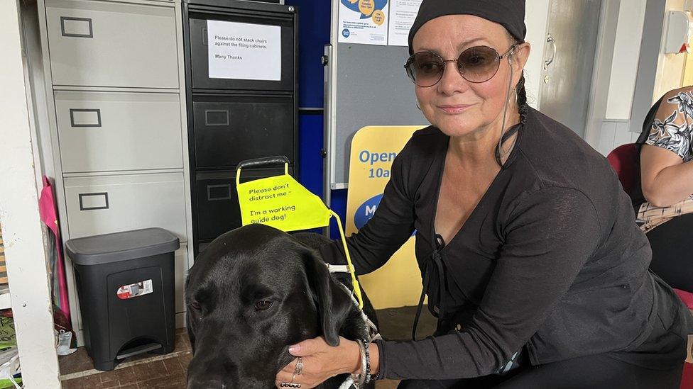 a woman dressed in black with dark glasses and her black Labrador guide dog