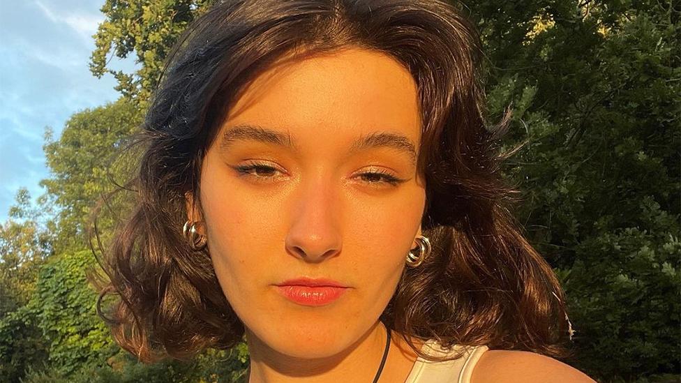 Valeria, a Ukrainian woman, looking at the camera, with the sun shining on her face. She is wearing two round hoop earrings, with the background of green trees.