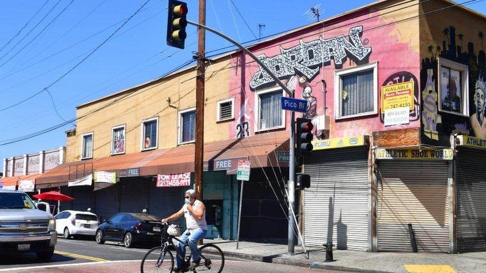 A man adjusts his facemask riding past closed shopfronts in what would normally be a busy fashion district in Los Angeles, California on May 4, 2020.