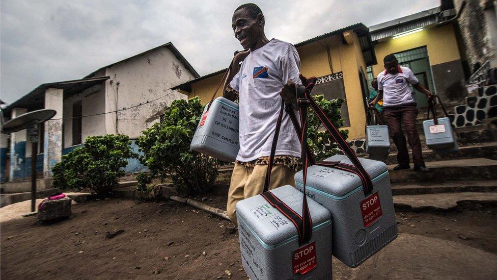 men carry cooler boxes of vaccines down the steps outside a building with a corrugated tin roof