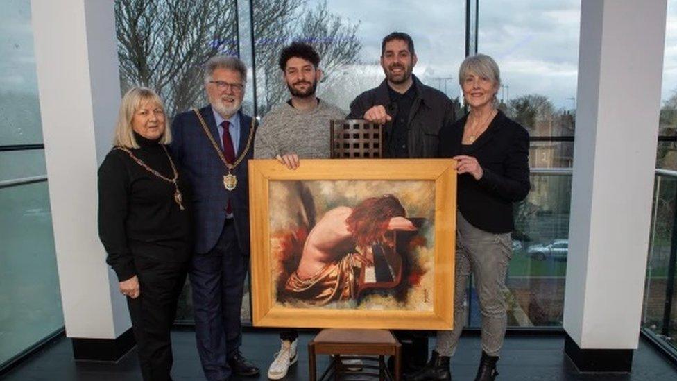 Two people wearing mayoral chains, two young men and a woman stand behind a painting