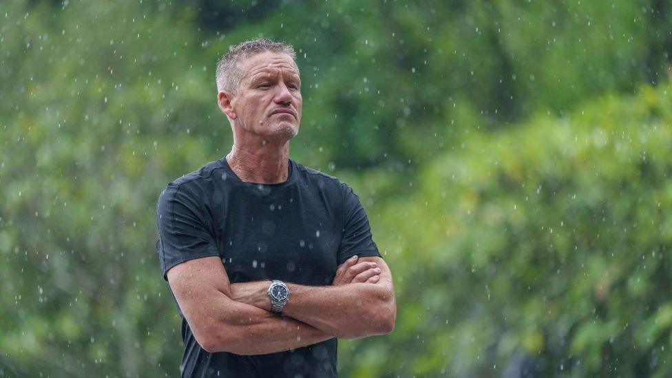 Billy Billingham is stood with his arms crossed in front of a background of trees. He has cropped hair and is wearing a black T-shirt.